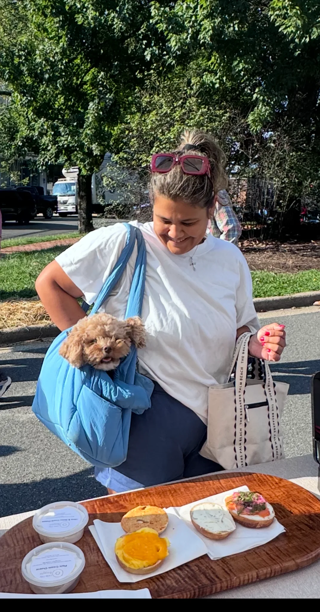 A smiling customer carrying a small dog in a blue bag beside a display of bagels and cream cheese at the market